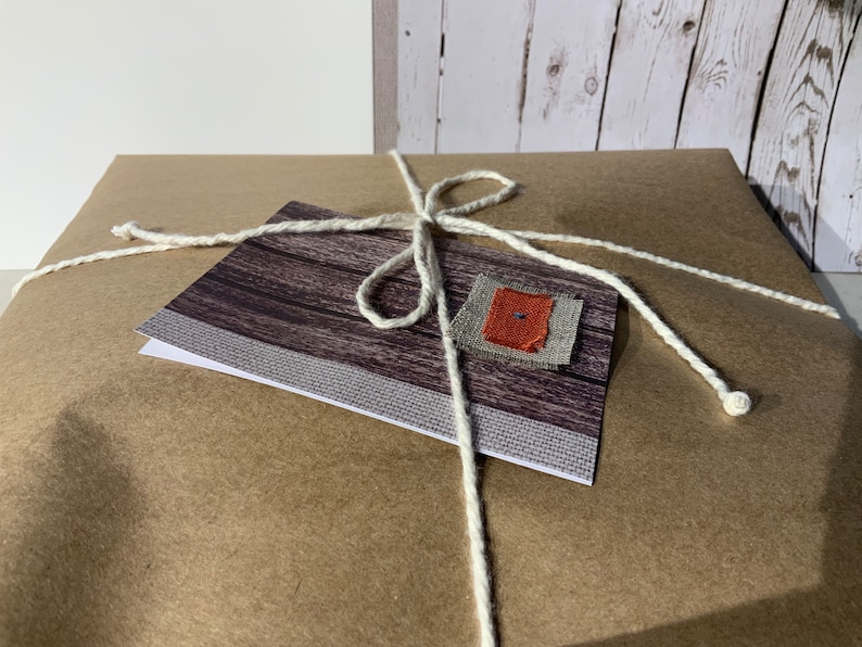 May include: A square package wrapped in brown paper and tied with a white string. A rectangular card with a dark wood grain pattern and a small square fabric patch in gray and orange is secured to the top of the package by the white string.
