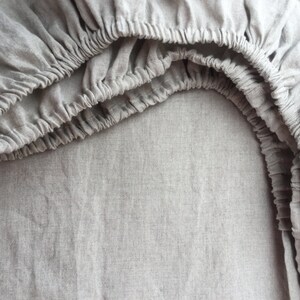 May include: A close-up view of a light gray fitted sheet. The elasticized edges are gathered, creating a textured, ruffled appearance. The fabric has a subtle, natural weave, suggesting a linen or similar material. The sheet is designed for a bed.
