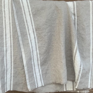 May include: A rectangular textile piece in a natural beige tone featuring vertical white stripes of varying widths. The fabric displays a visible woven texture with frayed edges along the bottom hem, creating a simple and functional design.