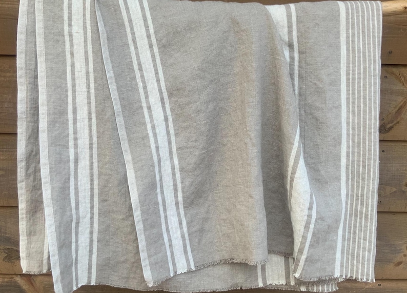 May include: A rectangular textile piece in a natural beige tone featuring vertical white stripes of varying widths. The fabric displays a visible woven texture with frayed edges along the bottom hem, creating a simple and functional design.