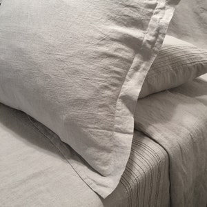 May include: Close-up of a light gray linen bedding set. The set includes a pillow with a detailed border and a pillowcase, along with a matching sheet. The fabric has a textured appearance.