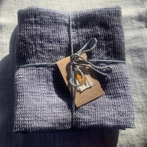 Puede incluir: Un textil cuadrado plegado con un patr&oacute;n de tejido de gofre en tonos azul oscuro y gris. La tela est&aacute; atada con una cuerda gris que asegura una peque&ntilde;a etiqueta de papel marr&oacute;n rectangular con un diminuto parche de tela amarillo y blanco cosido.