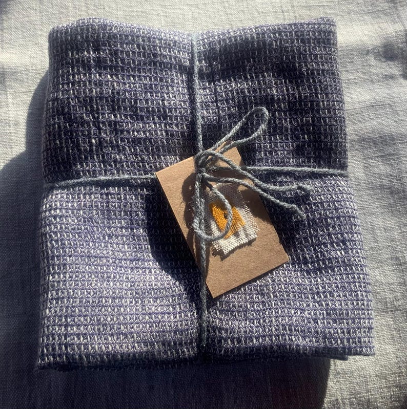 Puede incluir: Un textil cuadrado plegado con un patr&oacute;n de tejido de gofre en tonos azul oscuro y gris. La tela est&aacute; atada con una cuerda gris que asegura una peque&ntilde;a etiqueta de papel marr&oacute;n rectangular con un diminuto parche de tela amarillo y blanco cosido.