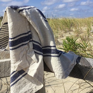 May include: A rectangular textile piece featuring a natural beige base color with prominent horizontal navy blue stripes. The fabric has a visible woven texture and is draped over a wooden structure, displaying a soft, folded appearance throughout.