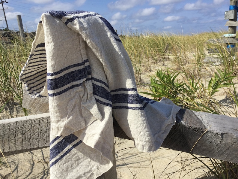 May include: A rectangular textile piece featuring a natural beige base color with prominent horizontal navy blue stripes. The fabric has a visible woven texture and is draped over a wooden structure, displaying a soft, folded appearance throughout.