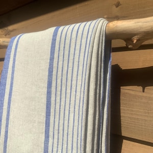 May include: A rectangular textile featuring a natural beige base colour with vertical blue stripes of varying widths. The fabric is draped over a light-coloured wooden branch, displaying a woven texture with visible threads throughout the material.