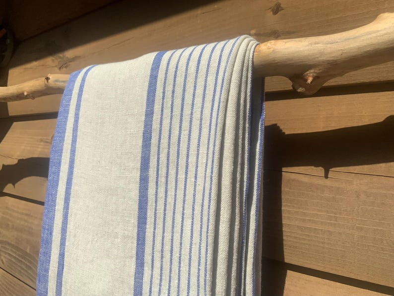 May include: A rectangular textile featuring a natural beige base colour with vertical blue stripes of varying widths. The fabric is draped over a light-coloured wooden branch, displaying a woven texture with visible threads throughout the material.
