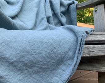May include: A soft, light blue textile throw blanket with a visible woven texture and natural folds, draped over the arm and seat of a weathered wooden chair. The fabric has a matte finish and a consistent, muted blue color throughout the piece.