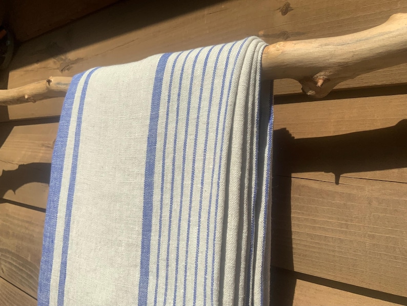 May include: A rectangular textile featuring a natural beige base color with vertical blue stripes of varying widths. The fabric is draped over a light-colored wooden branch, displaying a woven texture with visible threads throughout the material.