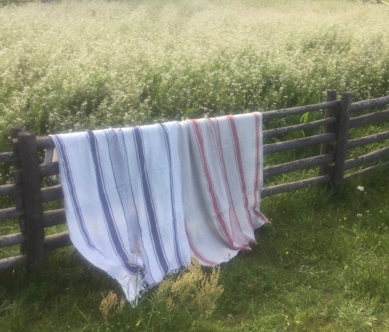 May include: Two rectangular woven textiles draped over a wooden fence. One textile features vertical blue stripes on a white background, while the other displays vertical red stripes on a white background. Both items have fringed edges at the bottom.