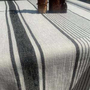 May include: A rectangular off-white linen tablecloth featuring a series of vertical black stripes of varying widths. The fabric has a visible woven texture, and the stripes are arranged in a repeating pattern across the surface of the textile.