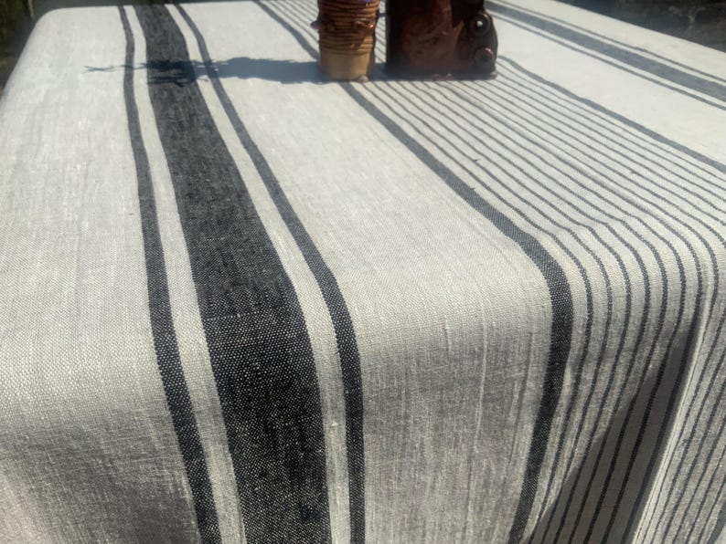 May include: A rectangular off-white linen tablecloth featuring a series of vertical black stripes of varying widths. The fabric has a visible woven texture, and the stripes are arranged in a repeating pattern across the surface of the textile.