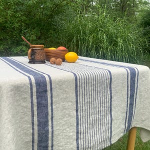May include: A linen tablecloth with blue stripes, set outdoors. A wooden bowl holds fruit, and a decorative mug and lemon sit on the table. The tablecloth is draped over a wooden table.