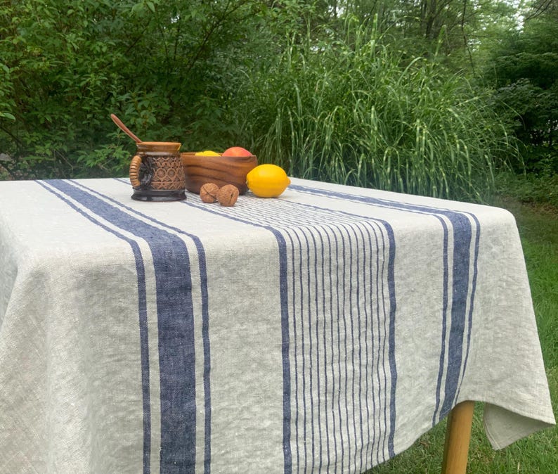 May include: A rectangular tablecloth made of light beige fabric featuring vertical navy blue stripes of varying widths. The tablecloth is draped over a table, with a brown ceramic mug, a wooden bowl containing fruit, and walnuts on top.