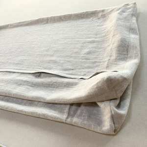 May include: A rectangular beige pillow cover made of a textured woven fabric. The cover features a visible zipper closure running along the side, with a folded corner showing the thickness and construction of the textile material.