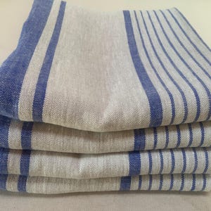May include: A stack of four folded rectangular napkins made of a natural beige fabric. Each napkin features a pattern of bold blue stripes along one side and a series of thinner, parallel blue pinstripes covering the remaining surface area.