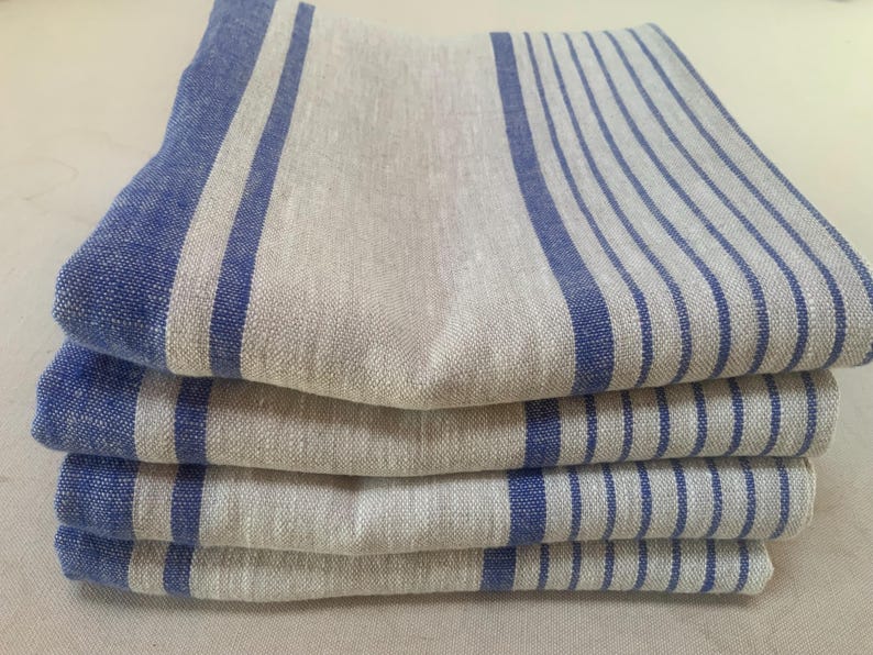May include: A stack of four folded rectangular napkins made of a natural beige fabric. Each napkin features a pattern of bold blue stripes along one side and a series of thinner, parallel blue pinstripes covering the remaining surface area.