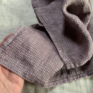 Puede incluir: Un tejido de color morado con textura de panal plegado que presenta un patr&oacute;n de cuadr&iacute;cula distintivo. La tela muestra un dise&ntilde;o cuadrado consistente en toda su superficie, exhibiendo un tono violeta apagado con sutiles variaciones.