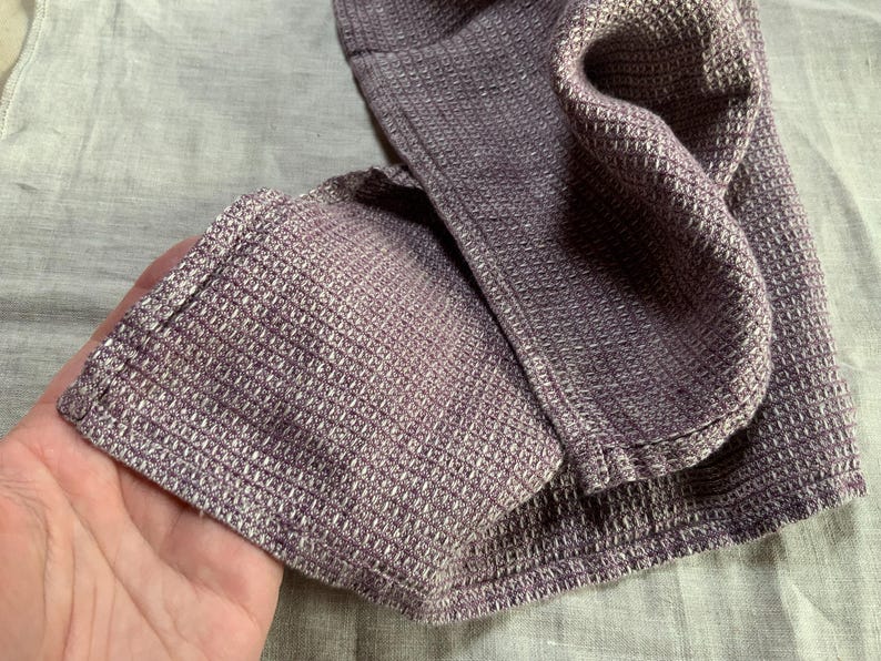 Puede incluir: Un tejido de color morado con textura de panal plegado que presenta un patr&oacute;n de cuadr&iacute;cula distintivo. La tela muestra un dise&ntilde;o cuadrado consistente en toda su superficie, exhibiendo un tono violeta apagado con sutiles variaciones.
