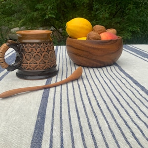 May include: A brown ceramic mug with a textured geometric pattern and a wrapped handle, a wooden bowl containing a yellow lemon, a peach, and walnuts, and a long, slender wooden spoon resting on a white and blue striped linen tablecloth.