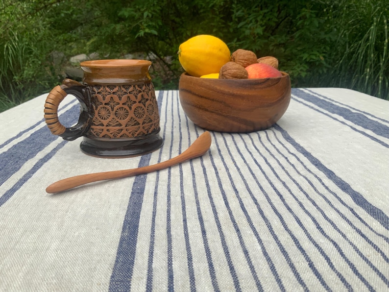 May include: A brown ceramic mug with a textured geometric pattern and a wrapped handle, a wooden bowl containing a yellow lemon, a peach, and walnuts, and a long, slender wooden spoon resting on a white and blue striped linen tablecloth.