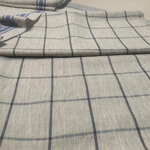 May include: A light beige linen fabric featuring a dark blue grid pattern of intersecting vertical and horizontal lines. The textile is folded to reveal a section with additional blue striped borders along the edge of the woven material.