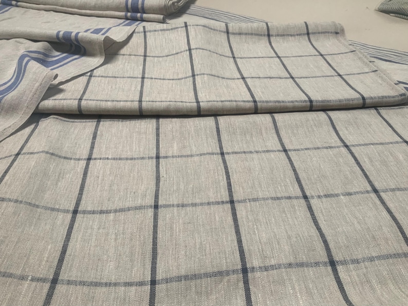 May include: A light beige linen fabric featuring a dark blue grid pattern of intersecting vertical and horizontal lines. The textile is folded to reveal a section with additional blue striped borders along the edge of the woven material.