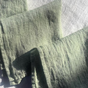 Puede incluir: Tres pa&ntilde;os de cocina de color verde salvia con una textura de tejido de gofre. La tela presenta un patr&oacute;n de cuadr&iacute;cula en toda su superficie, creando un dise&ntilde;o en relieve de cuadrados. Los pa&ntilde;os est&aacute;n doblados y superpuestos uniformemente.