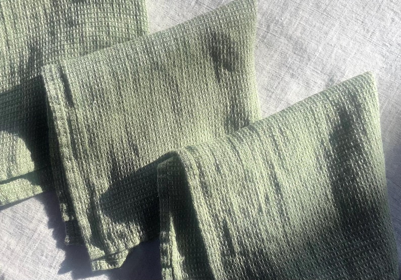 Puede incluir: Tres pa&ntilde;os de cocina de color verde salvia con una textura de tejido de gofre. La tela presenta un patr&oacute;n de cuadr&iacute;cula en toda su superficie, creando un dise&ntilde;o en relieve de cuadrados. Los pa&ntilde;os est&aacute;n doblados y superpuestos uniformemente.