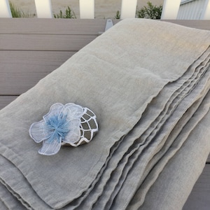 May include: A folded beige linen fabric piece featuring a decorative stone accent. The stone is wrapped in white crocheted netting and topped with a light blue fabric flower. The fabric has a visible weave and finished edges, creating a layered look.