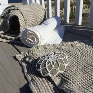 May include: A set of three waffle weave linen throw blankets in natural and white colors. The blankets are rolled up and arranged on a wooden deck. A white crocheted doily is placed on top of one of the blankets.