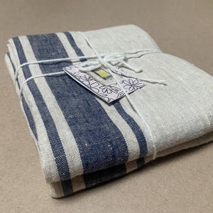 May include: A folded rectangular textile featuring a natural beige base with thick navy blue vertical stripes. The fabric is tied with white string and includes a small white tag with a black floral outline and a yellow rectangular accent.