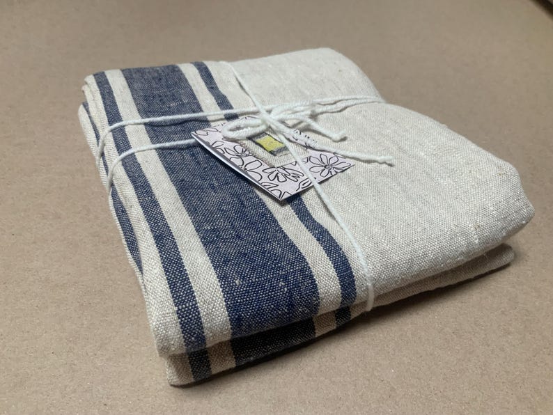 May include: A folded rectangular textile featuring a natural beige base with thick navy blue vertical stripes. The fabric is tied with white string and includes a small white tag with a black floral outline and a yellow rectangular accent.