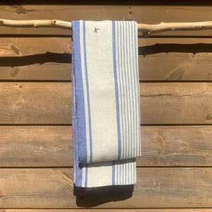 May include: A rectangular kitchen towel featuring a light beige base with vertical blue stripes of varying widths. The fabric displays a small, dark embroidered bird icon near the top edge, and the towel is folded over a horizontal wooden branch.