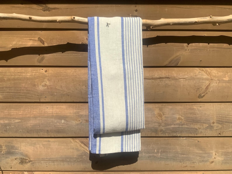 May include: A rectangular kitchen towel featuring a light beige base with vertical blue stripes of varying widths. The fabric displays a small, dark embroidered bird icon near the top edge, and the towel is folded over a horizontal wooden branch.