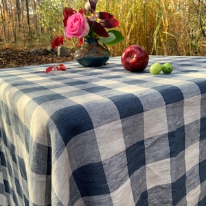 May include: A rectangular tablecloth featuring a large-scale navy blue and off-white buffalo check pattern. The fabric has a visible woven texture. The tablecloth is draped over a surface, creating soft folds along the edges and corners.