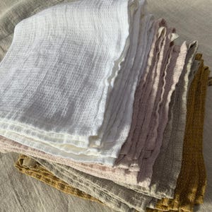 Puede incluir: Una pila de pa&ntilde;os plegados con tejido de gofre en una variedad de colores que incluyen blanco, rosa claro, malva, beige y amarillo mostaza. Cada pa&ntilde;o presenta una textura de rejilla distintiva y bordes suaves y deshilachados, apilados ordenadamente.
