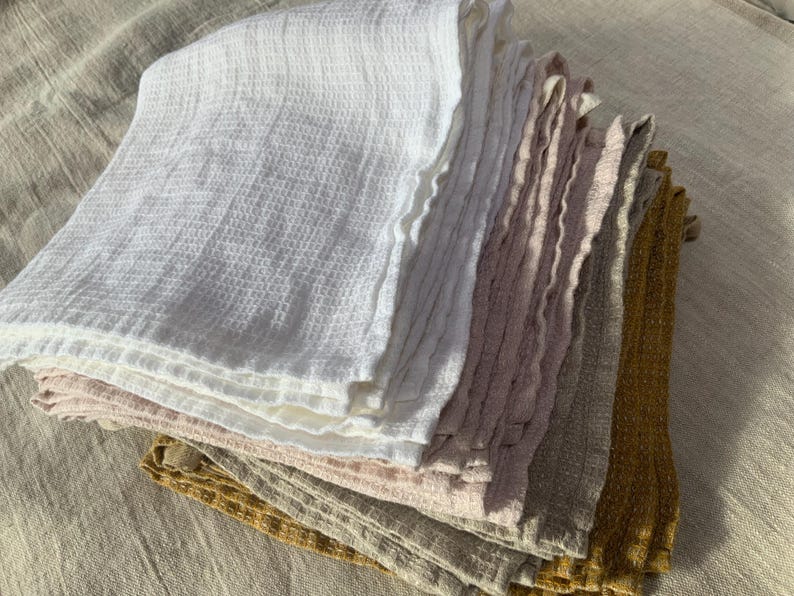 Puede incluir: Una pila de pa&ntilde;os plegados con tejido de gofre en una variedad de colores que incluyen blanco, rosa claro, malva, beige y amarillo mostaza. Cada pa&ntilde;o presenta una textura de rejilla distintiva y bordes suaves y deshilachados, apilados ordenadamente.