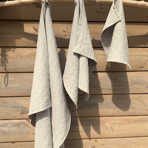 May include: Three light beige, rectangular kitchen towels of varying lengths are suspended from a horizontal wooden branch. Each towel features a simple, woven texture and a plain, neutral color, hanging vertically with soft, natural folds.
