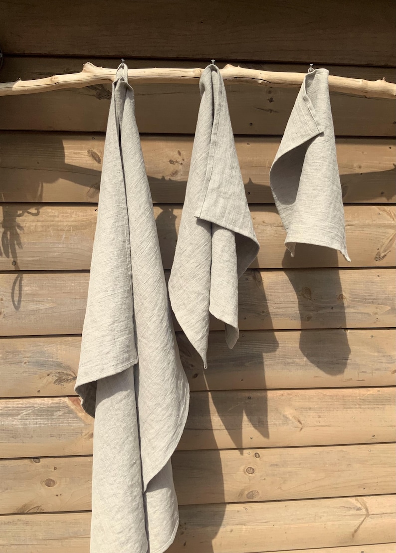 May include: Three light beige, rectangular kitchen towels of varying lengths are suspended from a horizontal wooden branch. Each towel features a simple, woven texture and a plain, neutral color, hanging vertically with soft, natural folds.