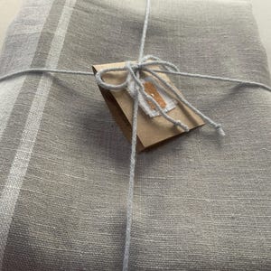 May include: A folded light grey textile with a visible woven texture and a vertical white stripe pattern. The fabric is tied with thin light grey string and features a small brown rectangular paper tag attached to the centre with a white label.