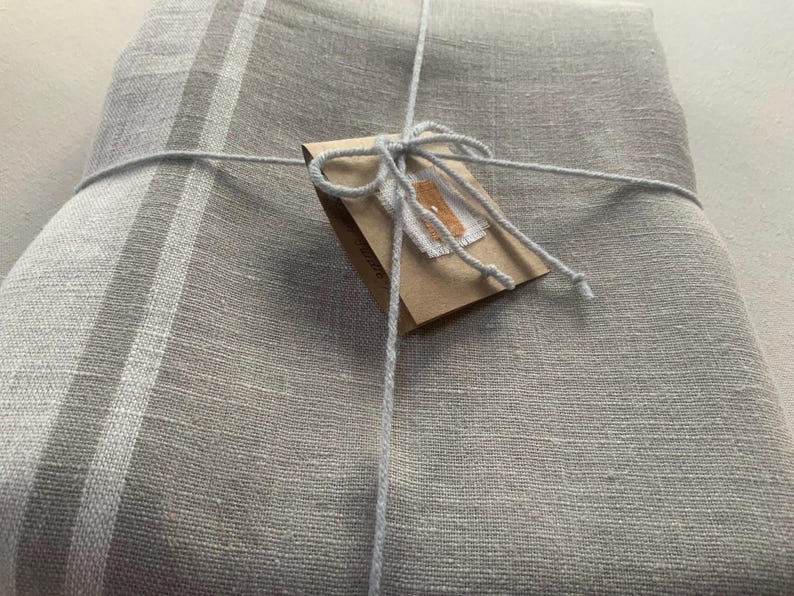 May include: A folded light grey textile with a visible woven texture and a vertical white stripe pattern. The fabric is tied with thin light grey string and features a small brown rectangular paper tag attached to the centre with a white label.