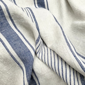May include: A piece of natural beige fabric featuring a pattern of bold navy blue stripes and thinner navy blue pinstripes. The textile has a visible woven texture and is draped to create soft folds, highlighting the contrast between the colors.
