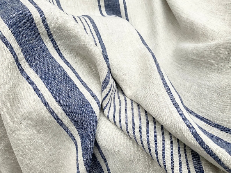 May include: A piece of natural beige fabric featuring a pattern of bold navy blue stripes and thinner navy blue pinstripes. The textile has a visible woven texture and is draped to create soft folds, highlighting the contrast between the colors.