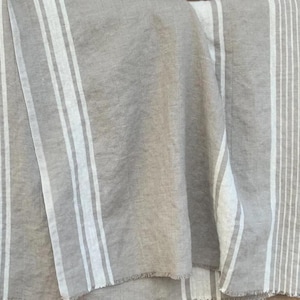 May include: A woven fabric textile featuring a neutral beige and off-white color palette. The material displays a pattern of vertical stripes of varying widths, with a visible textured weave and frayed edges along the bottom hem of the cloth.