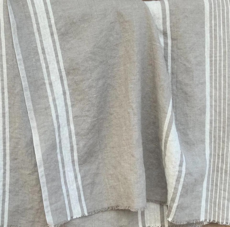 May include: A woven fabric textile featuring a neutral beige and off-white color palette. The material displays a pattern of vertical stripes of varying widths, with a visible textured weave and frayed edges along the bottom hem of the cloth.