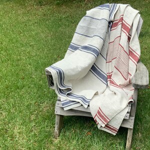 May include: A large, rectangular woven throw blanket featuring a cream-colored base with vertical stripes in shades of navy blue and deep red. The fabric has a visible, textured weave and is draped over a weathered wooden chair, showing striped patterns.