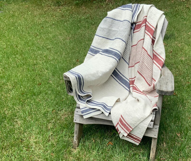 May include: A large, rectangular woven throw blanket featuring a cream-colored base with vertical stripes in shades of navy blue and deep red. The fabric has a visible, textured weave and is draped over a weathered wooden chair, showing striped patterns.