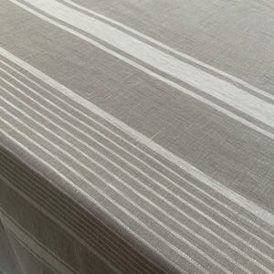 May include: A rectangular tablecloth made of a woven fabric in a neutral taupe and beige colour palette. The surface features a repeating pattern of wide and narrow vertical stripes in varying shades of off-white, creating a linear design.