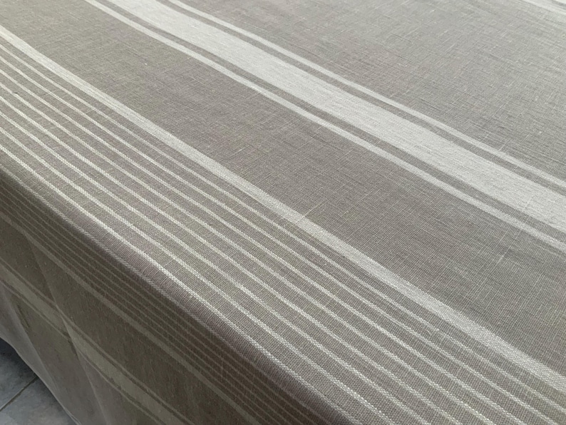 May include: A rectangular tablecloth made of a woven fabric in a neutral taupe and beige colour palette. The surface features a repeating pattern of wide and narrow vertical stripes in varying shades of off-white, creating a linear design.